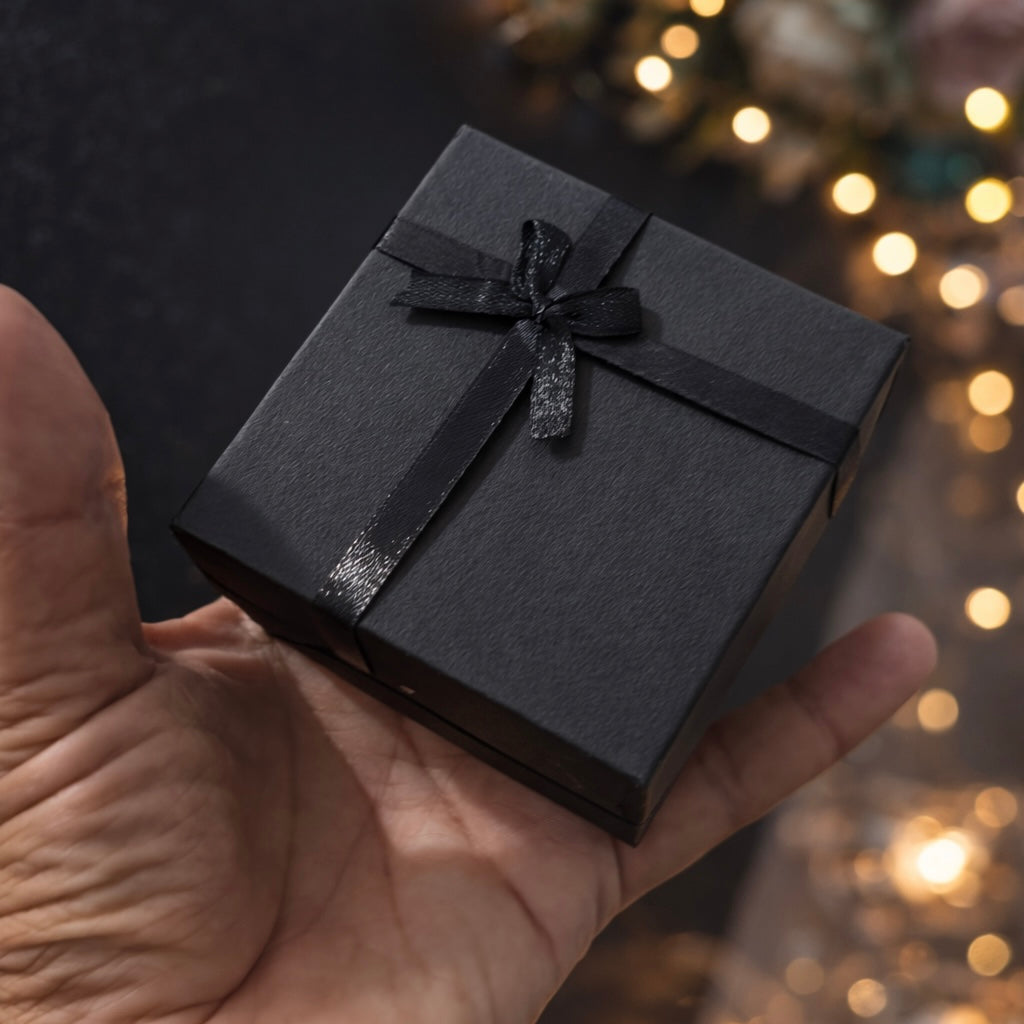 A small black gift box tied with a black ribbon and bow, held in a person's hand against a dark background with warm bokeh lights and blurred flowers.