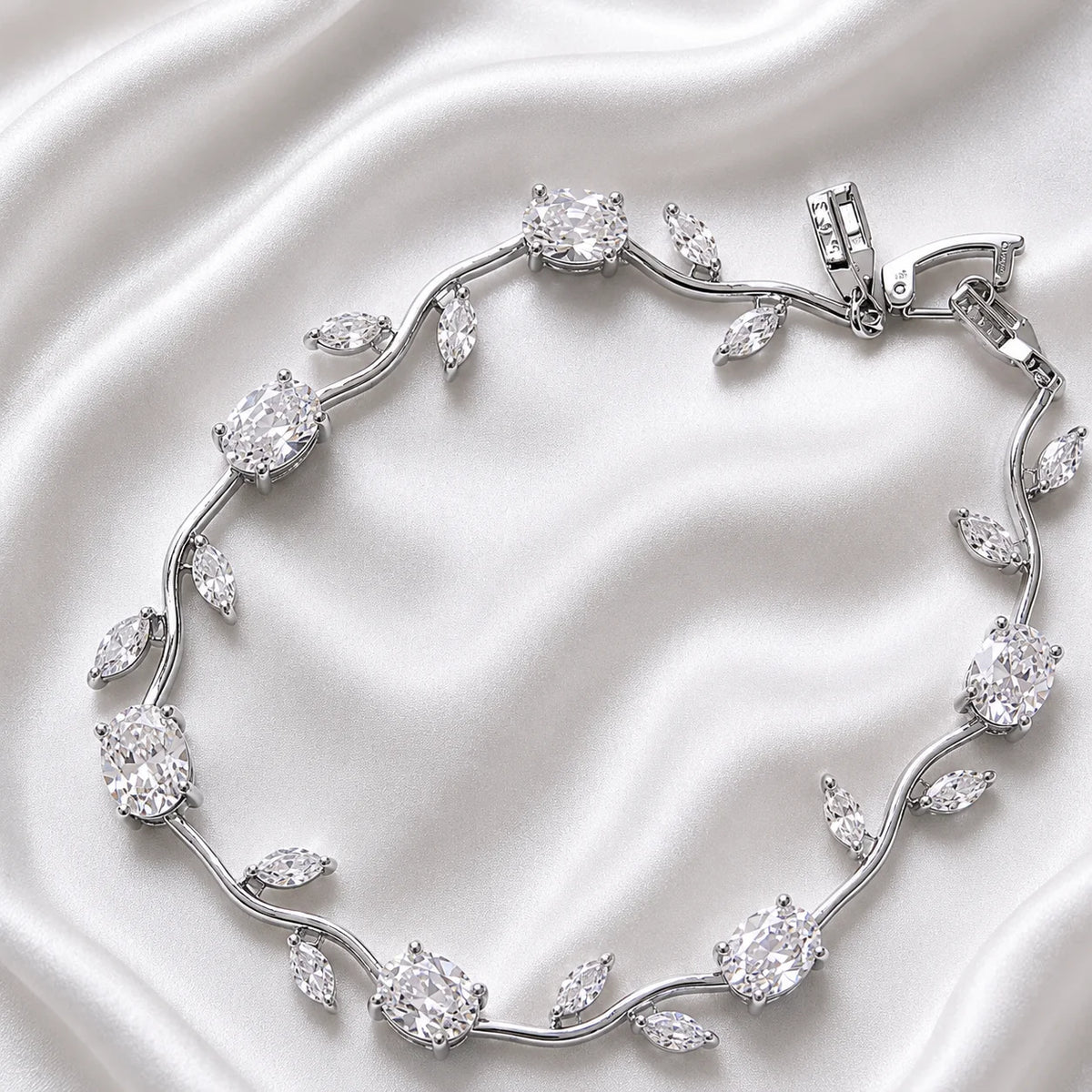 A silver-toned vine bracelet with clear gemstones arranged in leaf and oval motifs, photographed on a draped white satin fabric background.