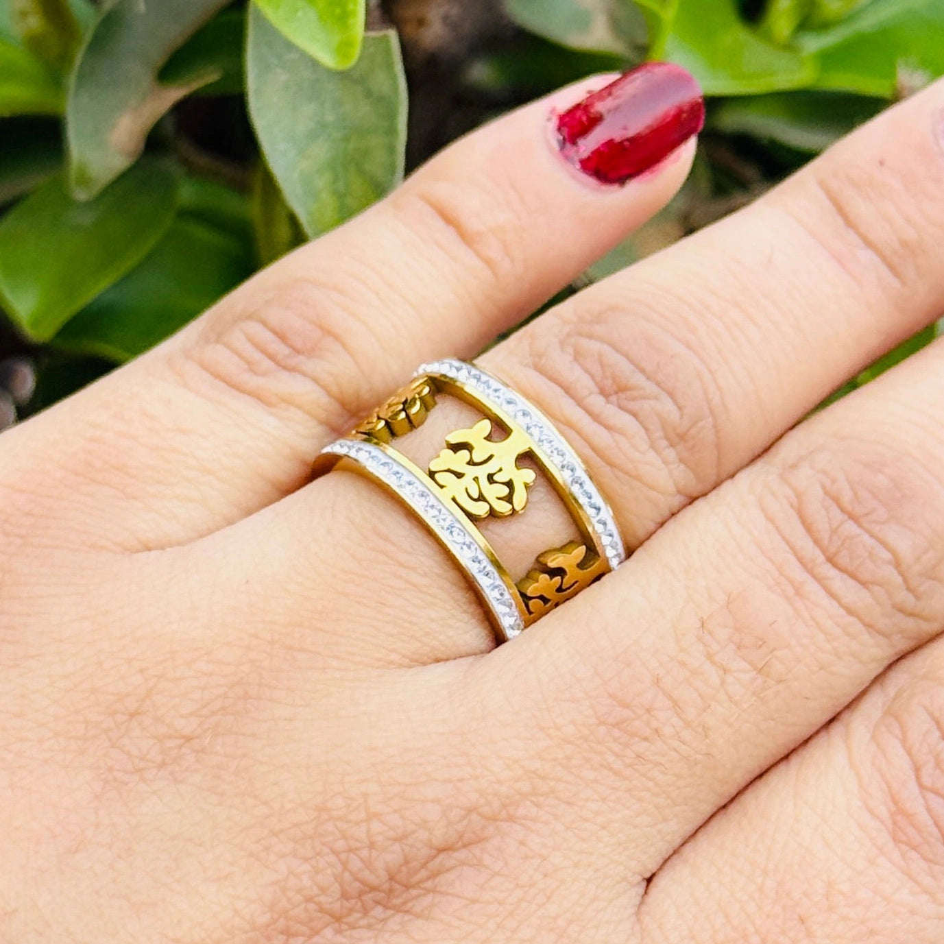 Hand wearing a gold ring with intricate design against a green leafy background