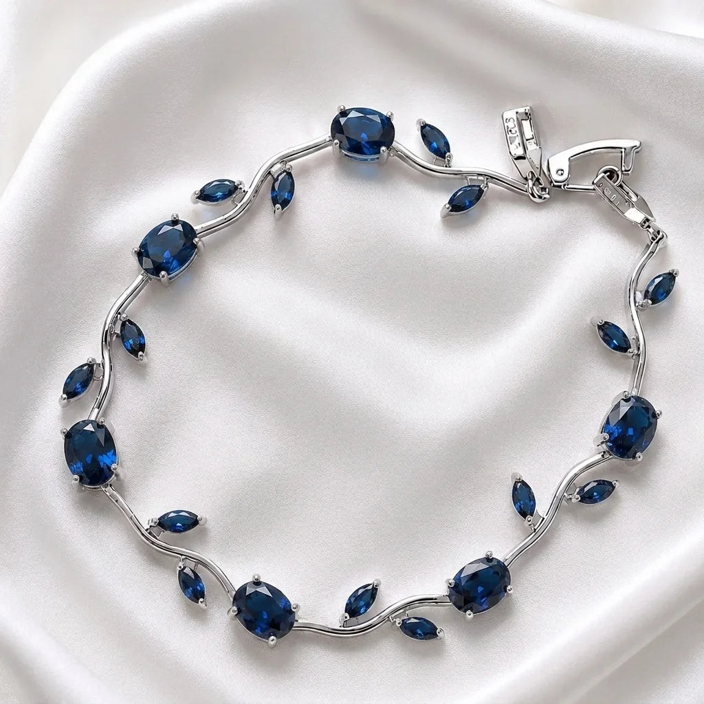 A silver vine-style bracelet featuring oval and marquise-cut deep blue gemstones, displayed on a soft white satin fabric background.