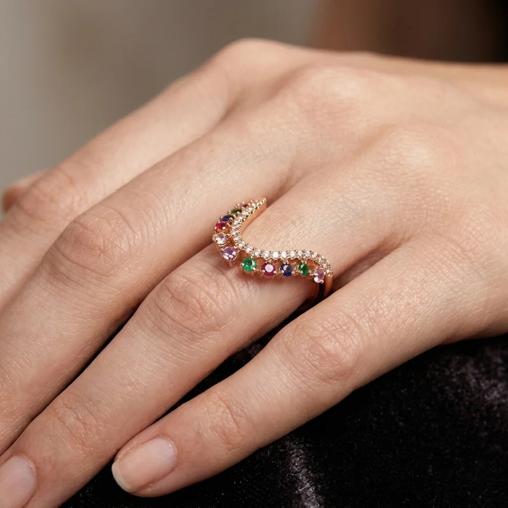 A close-up of a hand wearing a wavy rose gold ring adorned with small clear stones and a row of colorful gemstones including red, blue, green, and purple.