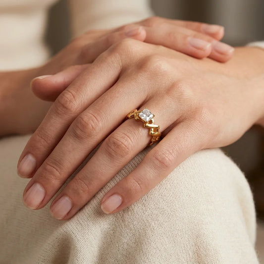 A close-up of a person's hands resting on their lap, wearing a gold ring with a clear rectangular gemstone and geometric gold details.