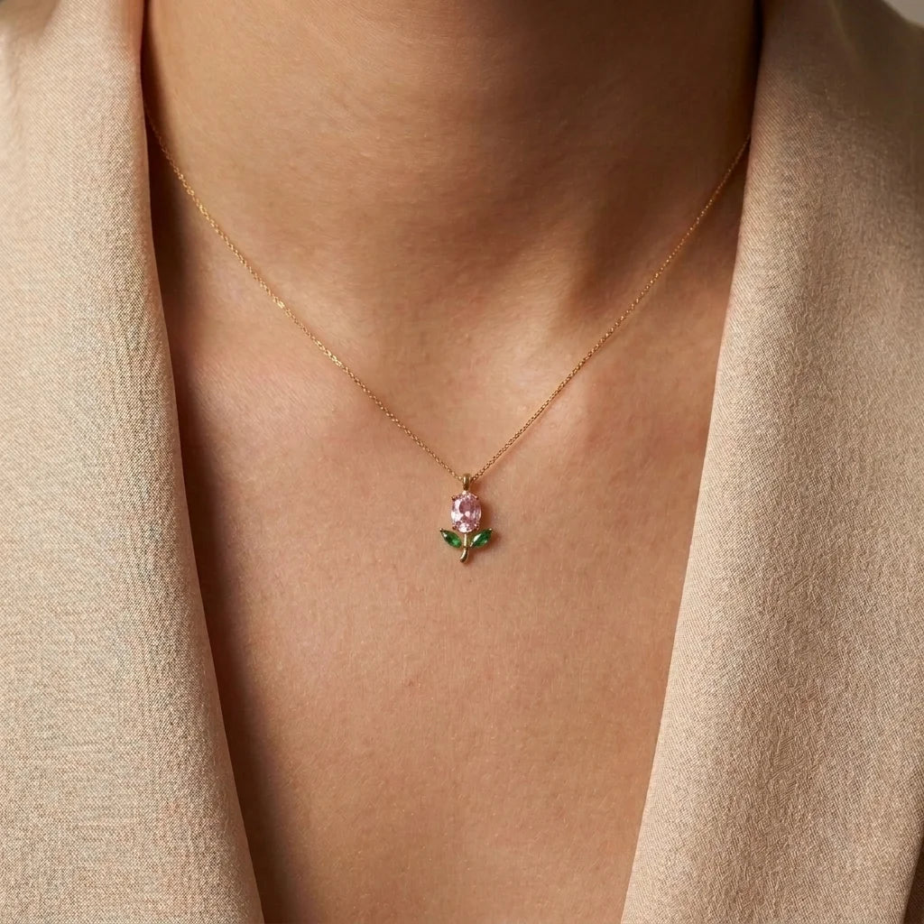 A close-up of a person wearing a delicate gold necklace with a small pendant featuring a pink flower and green leaves, styled with a beige blazer.