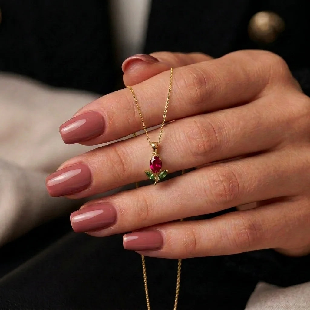 A close-up of a hand with mauve manicured nails holding a delicate gold chain necklace with a small pendant featuring a red gemstone and green leaf details.