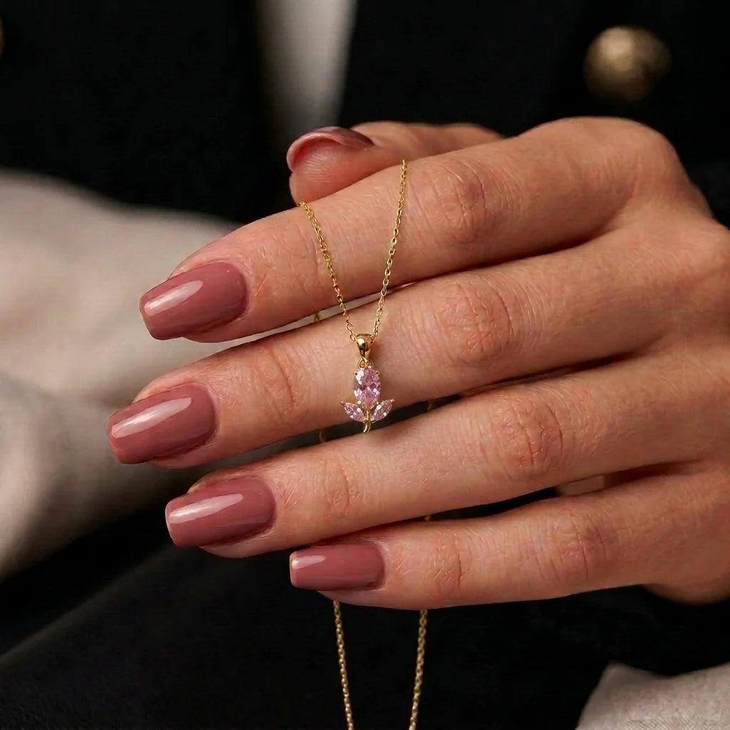 A close-up of a hand with mauve manicured nails holding a delicate gold chain necklace with a small pink teardrop gemstone pendant.