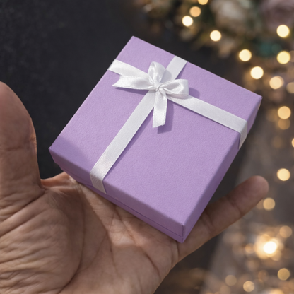 A small lavender gift box tied with a white ribbon and bow, held in a person's hand against a dark background with warm bokeh lights and blurred flowers.