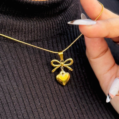 Gold necklace with a heart and bow pendant held by a hand against a black background