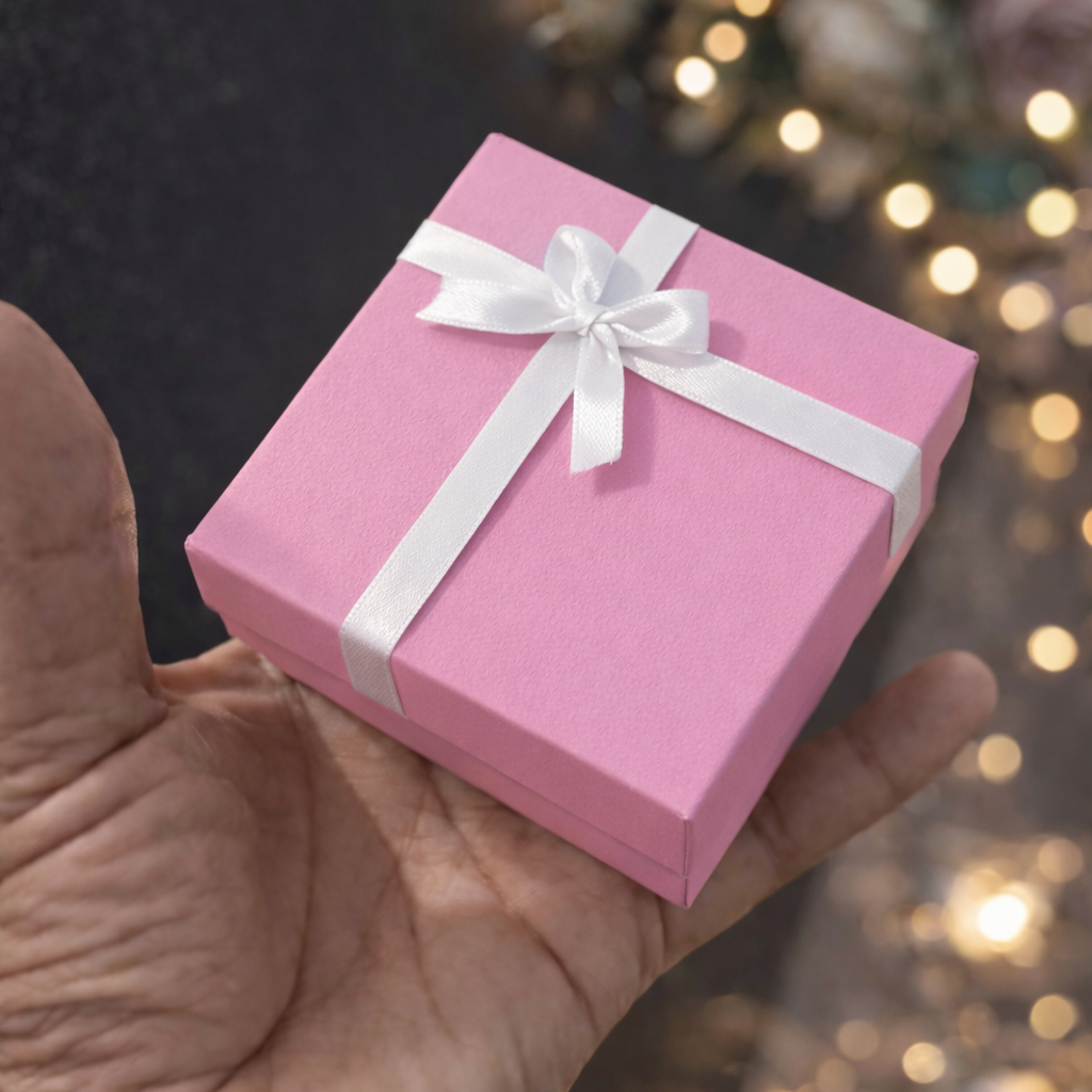 A small pink gift box tied with a white ribbon and bow, held in a person's hand against a dark background with warm bokeh lights.