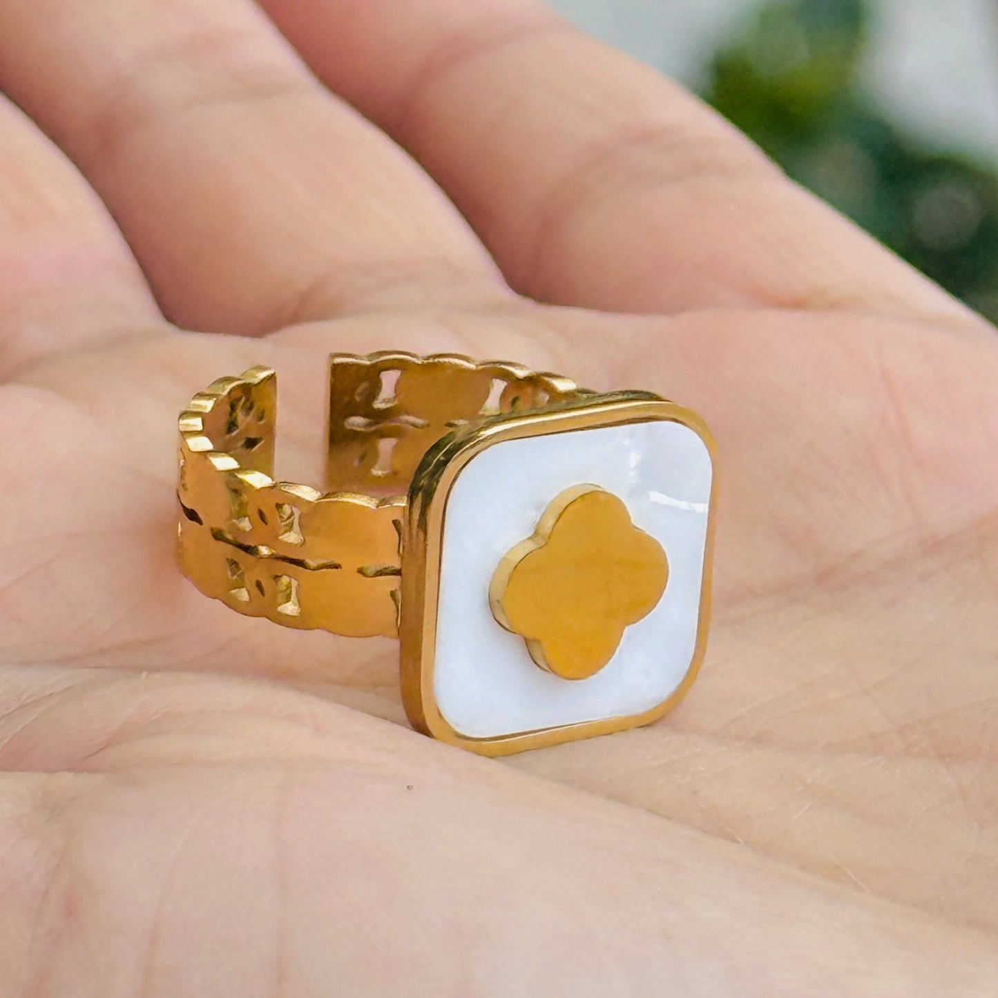 Gold ring with a white square and gold quatrefoil design on a hand.