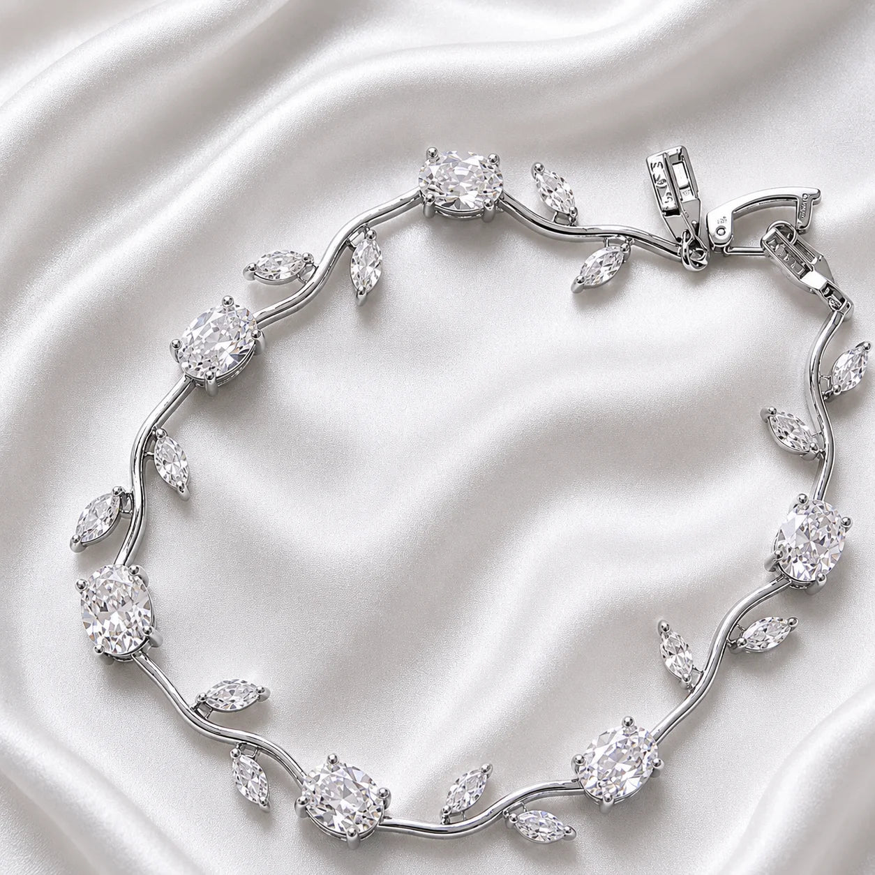 A silver-toned vine bracelet with clear gemstones arranged in leaf and oval motifs, photographed on a draped white satin fabric background.