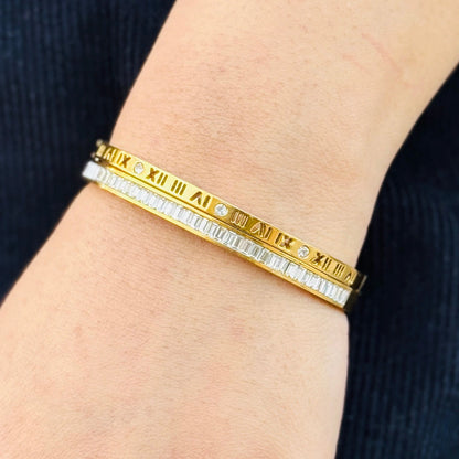 Gold bracelet with Roman numeral design on a wrist against a dark background