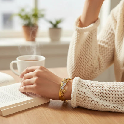 Person reading a book with a cup of coffee on a wooden table, wearing a beige sweater and gold bracelet.