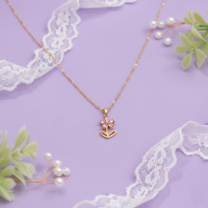 Gold necklace with a pink flower pendant on a purple background with lace and greenery.