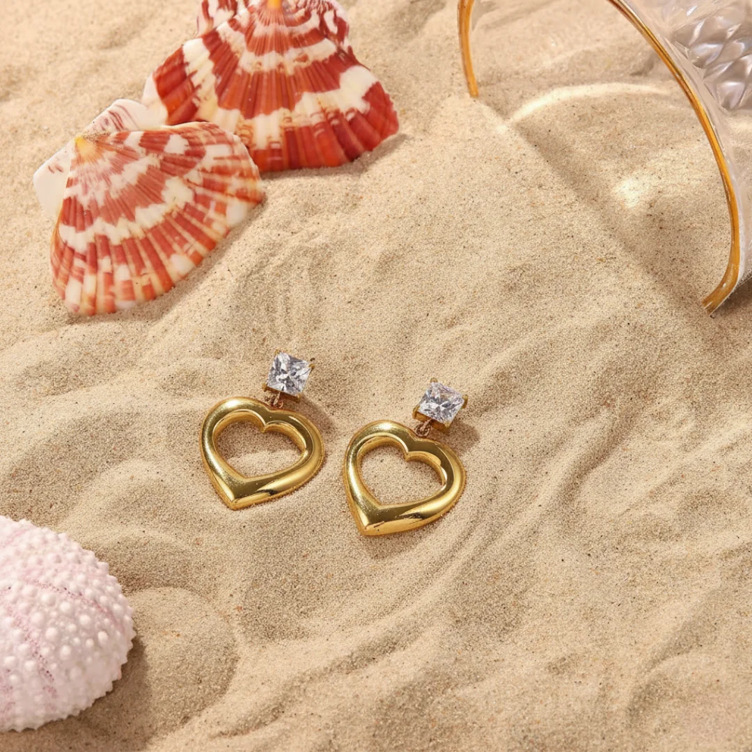 Gold heart-shaped earrings with clear stones on a sandy surface with shells and a glass of water.
