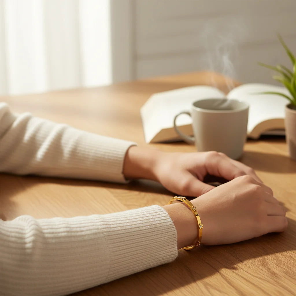 Person wearing a white sweater and gold bracelet on a wooden table with a cup and book in the background.