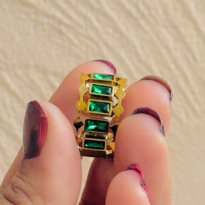 Gold ring with green gemstones held between fingers against a beige background