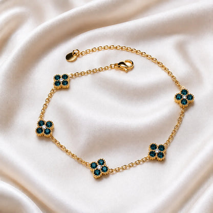 A gold chain bracelet with five floral clover motifs, each set with dark teal and clear rhinestones, displayed on a draped cream satin fabric.