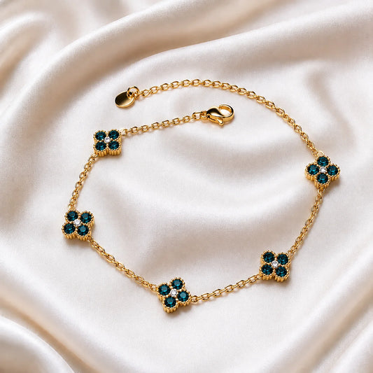 A gold chain bracelet with five floral clover motifs, each set with dark teal and clear rhinestones, displayed on a draped cream satin fabric.
