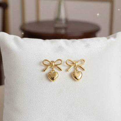 Gold bow-shaped earrings on a white pillow with a blurred background of a red rose in a vase.