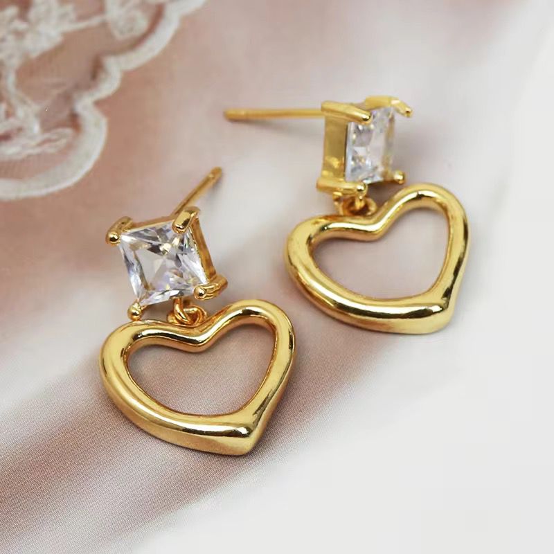 Gold heart-shaped earrings with clear gemstones on a soft pink background