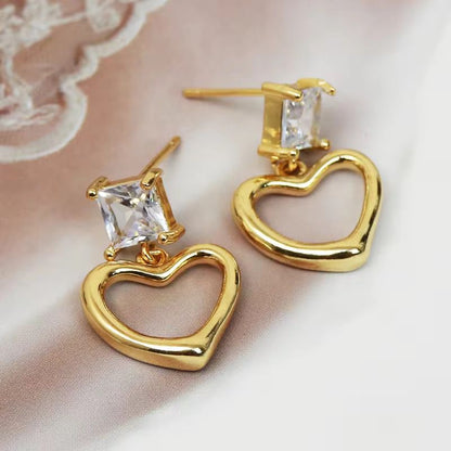 Gold heart-shaped earrings with clear gemstones on a soft pink background