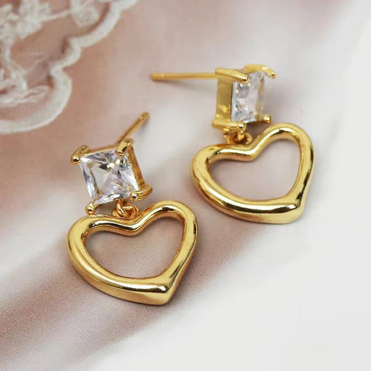 Gold heart-shaped earrings with clear gemstones on a soft pink background
