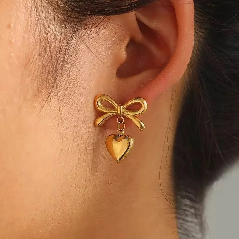 Gold bow and heart earring worn on an ear with a blurred background
