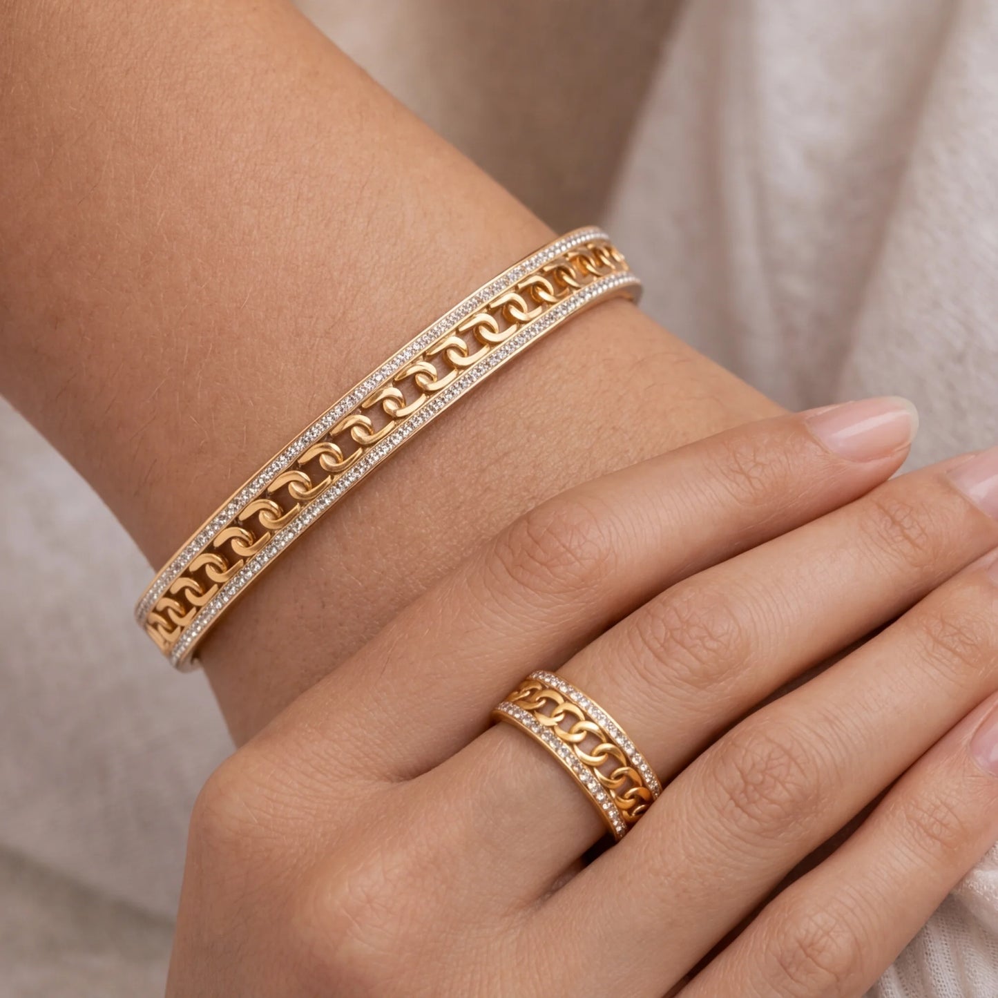 Gold chain bracelet and ring on a wrist against a neutral background