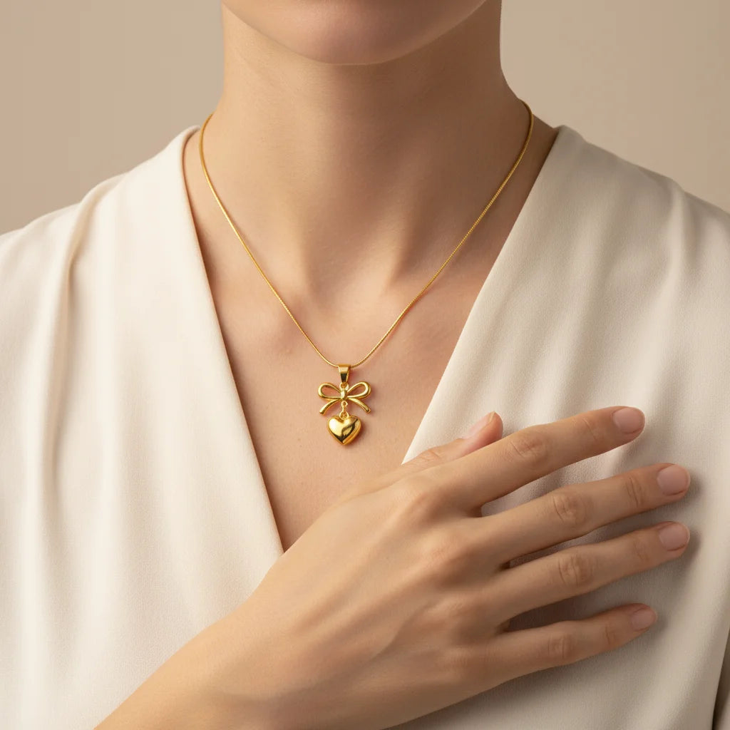 Gold necklace with a heart and bow pendant worn by a person in a white outfit.