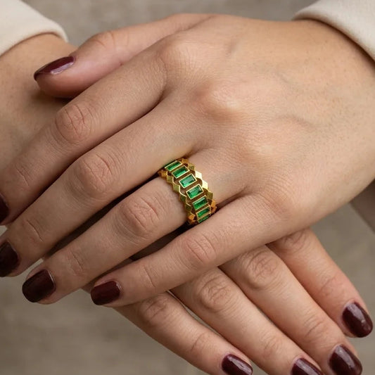 Close-up of a hand wearing a gold ring with green gemstones against a neutral background