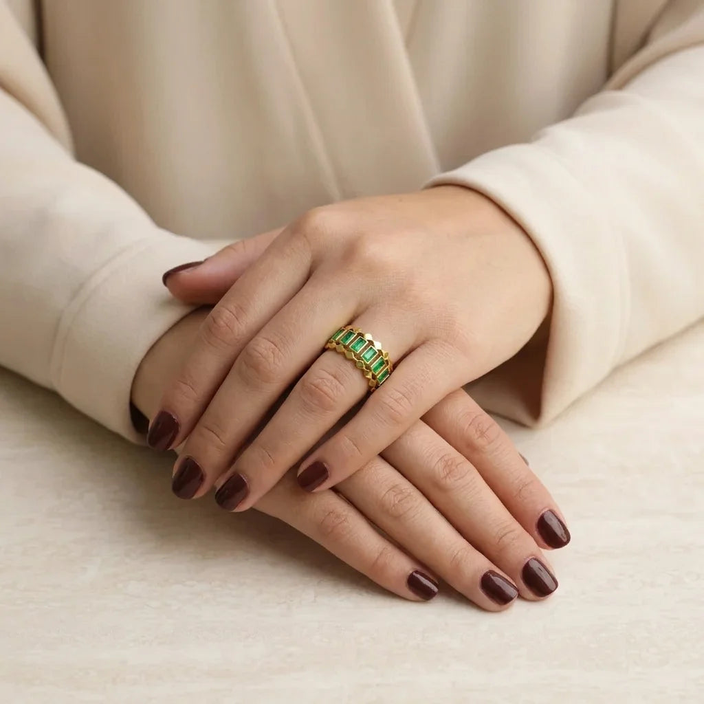 Hand wearing a gold ring with green gemstones on a beige background