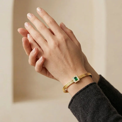 Hand wearing a gold bracelet with a green gemstone on a beige background