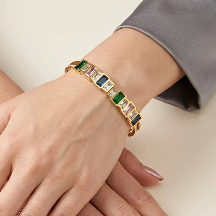 Gold bracelet with green and blue stones on a hand against a neutral background