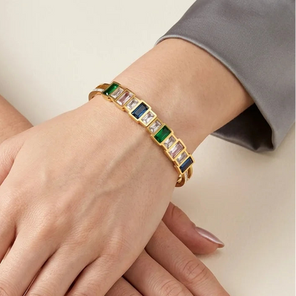 Gold bracelet with green and blue stones on a hand against a neutral background
