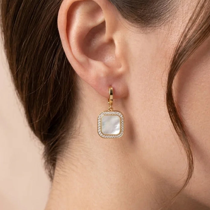 Close-up of a woman wearing gold earrings with a square gemstone on a neutral background