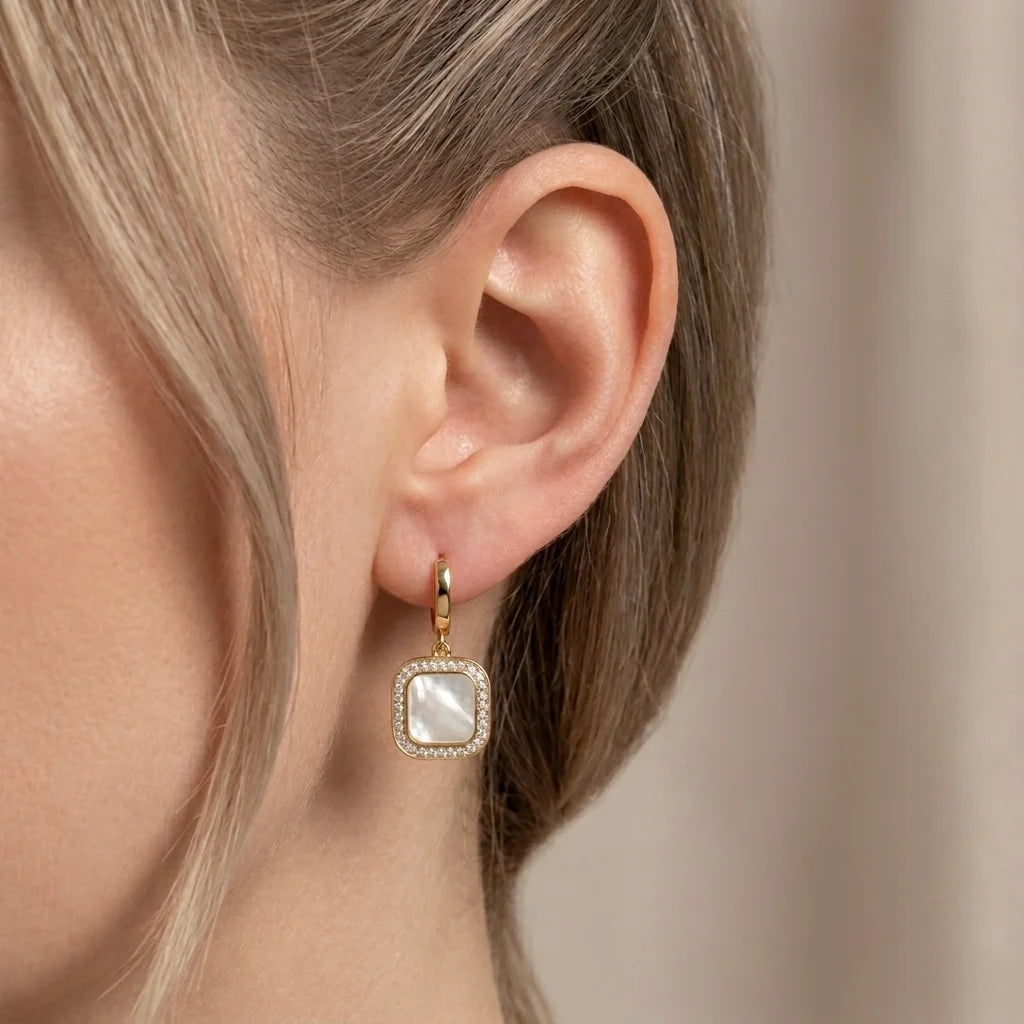 Close-up of an ear wearing a gold earring with a clear gemstone on a neutral background
