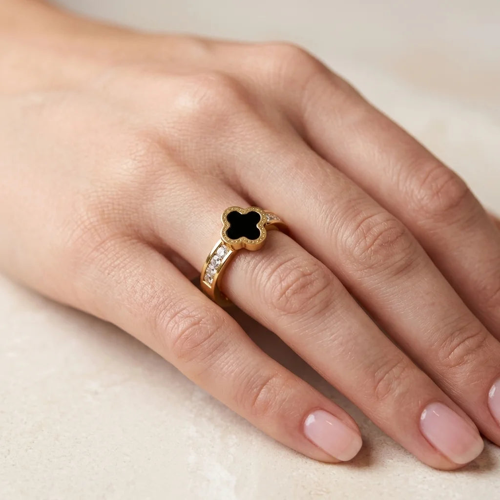 Gold ring with a black clover design on a hand against a neutral background