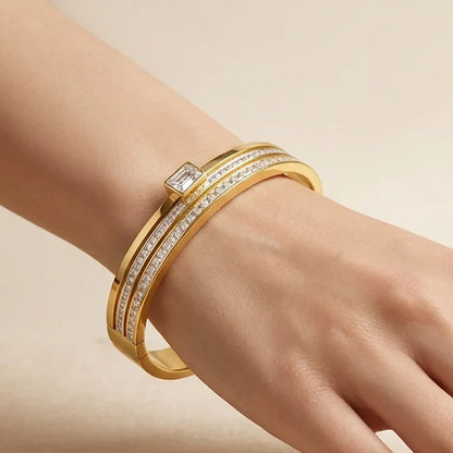 Hand wearing a gold bracelet with diamonds on a beige background, featuring the brand 'Aurelien'.