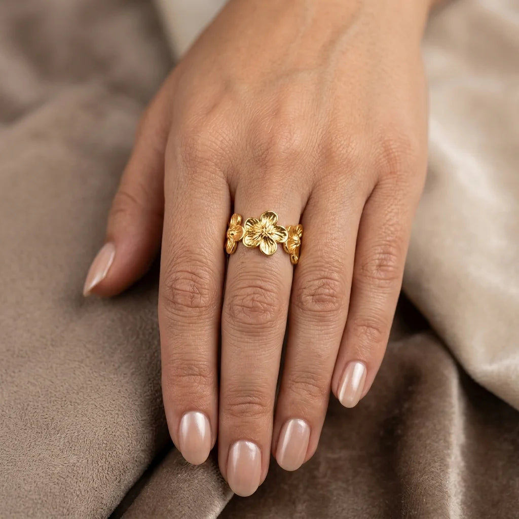 Gold floral ring on a hand with beige nail polish against a soft brown background