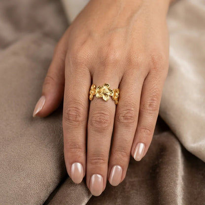 Gold floral ring on a hand with beige nail polish against a soft brown background