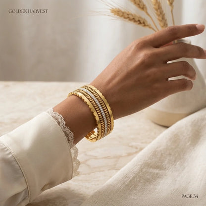 Hand wearing a gold bracelet with a blurred background featuring wheat stalks.