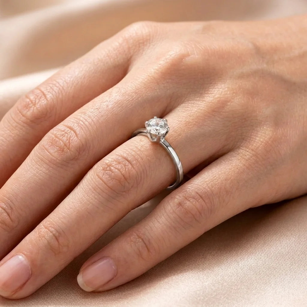 A silver solitaire engagement ring with a round-cut clear gemstone, shown worn on a person's ring finger against a soft beige satin background.