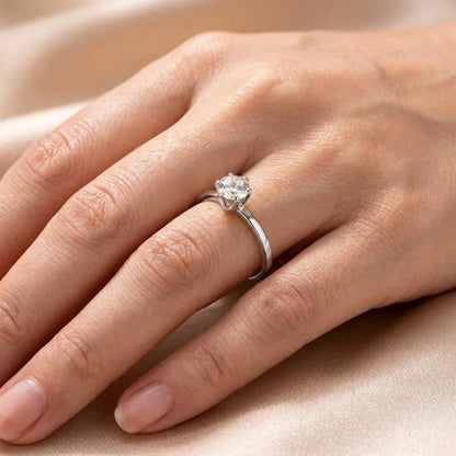 A silver solitaire engagement ring with a round-cut clear gemstone, shown worn on a person's ring finger against a soft beige satin background.