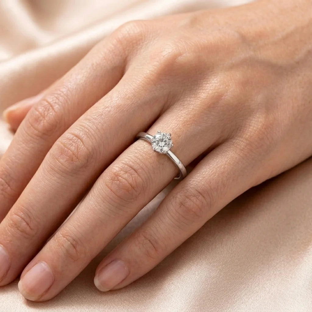A close-up of a hand wearing a silver-toned diamond engagement ring, resting on a soft beige satin fabric background.