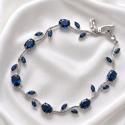A silver vine-style bracelet featuring oval and marquise-cut deep blue gemstones, displayed on a soft white satin fabric background.