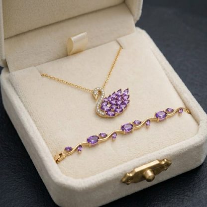 A gold jewelry set with a swan-shaped pendant necklace and a matching bracelet, both featuring purple gemstones, displayed inside an open cream-colored jewelry box.