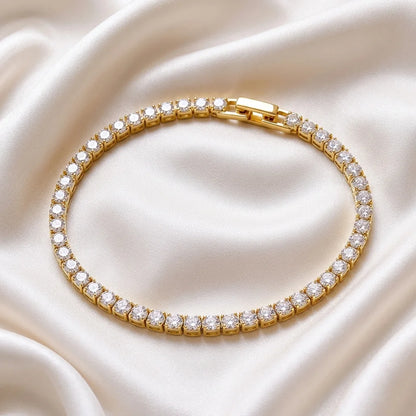 A gold tennis bracelet with clear stones, arranged in a circle on a draped cream satin fabric background.