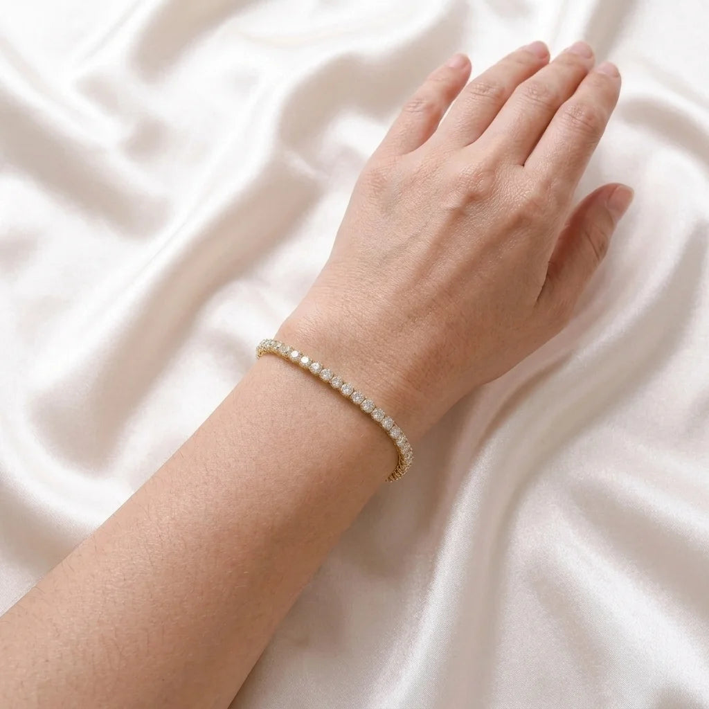 A delicate gold tennis bracelet with small clear stones worn on a person's wrist, resting against a draped white satin fabric background.