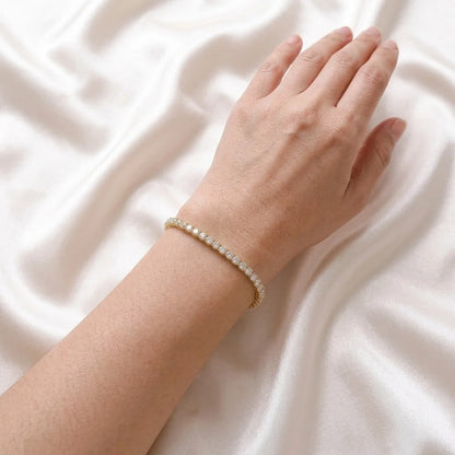 A delicate gold tennis bracelet with small clear stones worn on a person's wrist, resting against a draped white satin fabric background.