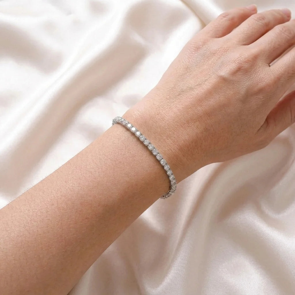 A delicate silver tennis bracelet with small round clear stones, worn on a person's wrist against a soft, draped white satin fabric background.